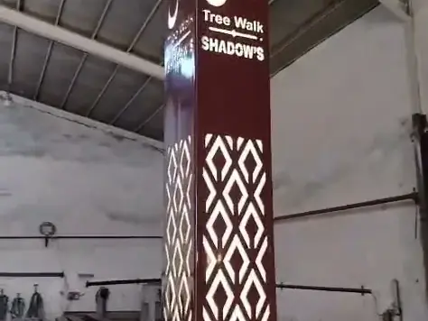 عمود إنارة ديكوري بقصّ ليزري بلون بنّي يحمل شعار مشروع Tree Walk، من تصنيع مصنع أكتار، مُضاء داخل المصنع